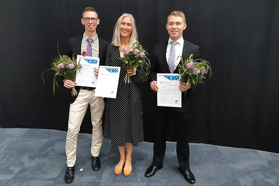 Tre personer, två män kavaj och i mitten på bilden en kvinna med svart- och vitprickig klänning, alla ler och håller varsitt stipendium.