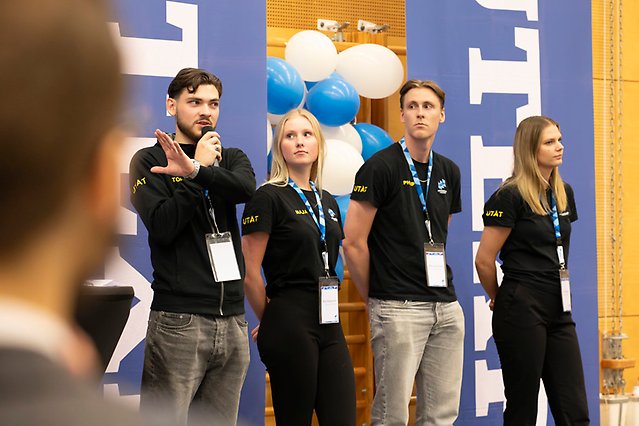 Fyra studenter i svarta t-shirts står på scen. Studenten längst till vänster talar i mikrofon och gestikulerar med handen. I bakgrunden finns ballonger. Foto.