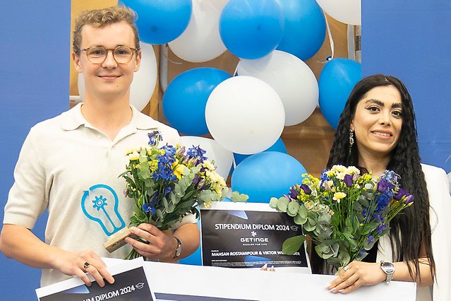 Två studenter håller i en varsin blombukett, statyett och check. Foto.