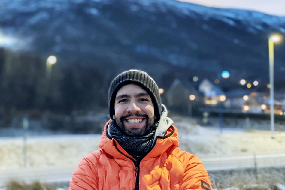 A man wearing a beanie, muffler and a jacket smiles at the camera. In the background is a mountain, with houses and lampposts beneath it. Photo.