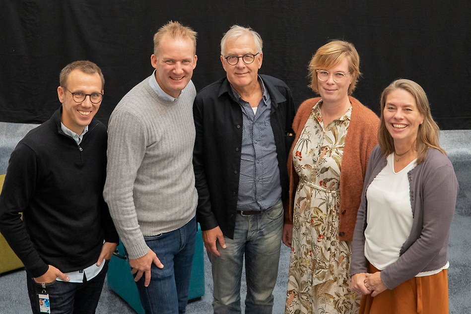 Fem personer i en liten halvcirkel, tittar mot kameran och ler.