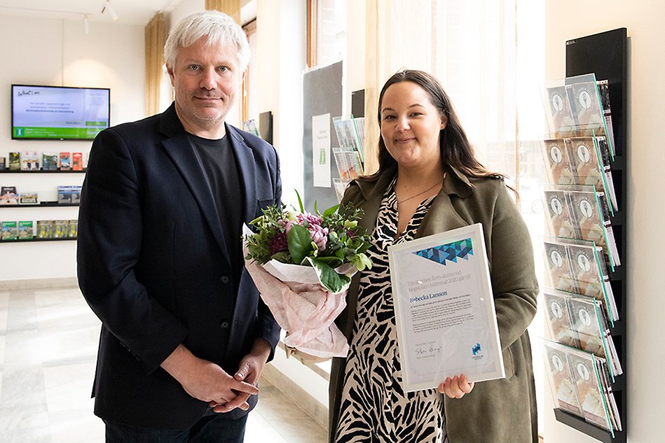 En kvinna står med blommor och diplom bredvid en man i kostym. Foto.