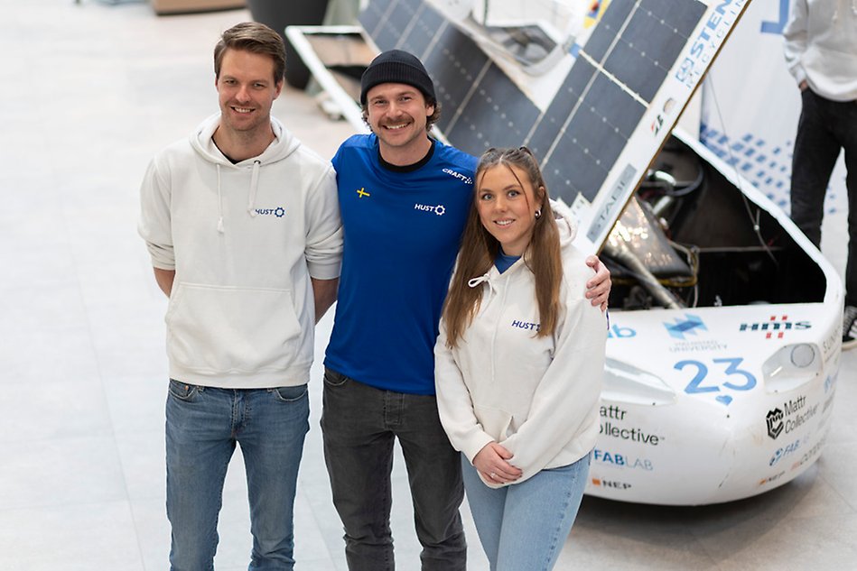 Two young men and a young woman are standing next to each other in a row. They are looking towards the camera. Behind them is a white racing car with solar panels on the roof.