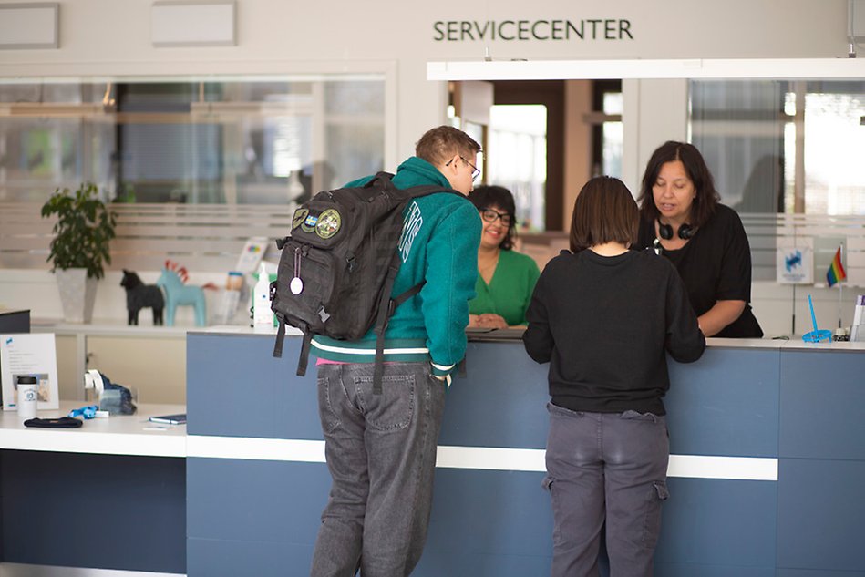 Tre personer står i servicecenter och pratar med en kvinna på andra sidan disken. Foto. 