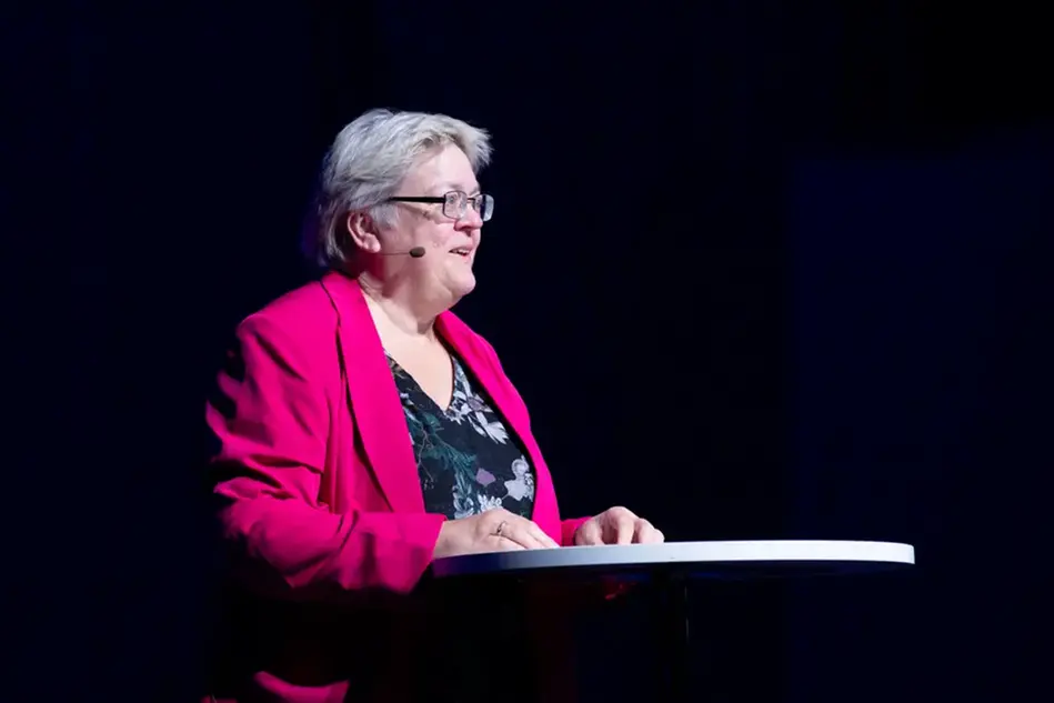 En kvinna med kort ljust hår och glasögon står vid ett podium. Foto.