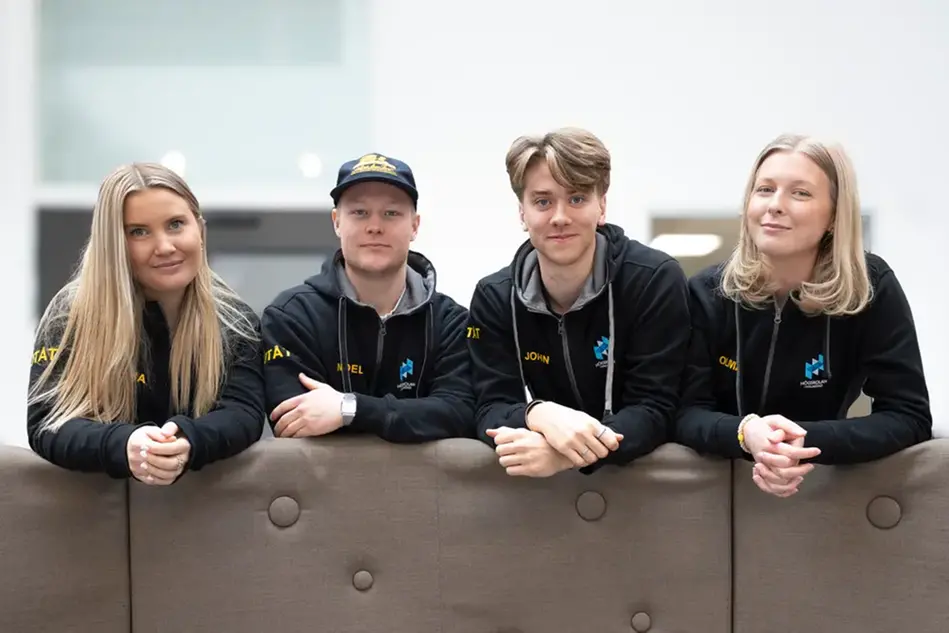 Four students in identical hoodies lean on an edge and smiles at the camera.