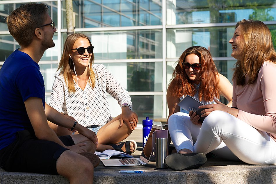 Fyra glada studenter sitter i skräddarställning på gräset i solen. I bakgrunden ses en stor glasvägg.