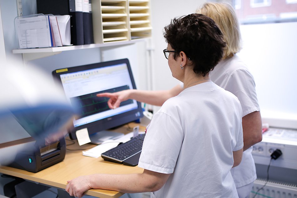 Två kvinnor i vårdkläder står framför en datorskärm i kontorsmiljö, den ena kvinnan pekar på skärmen. Foto.
