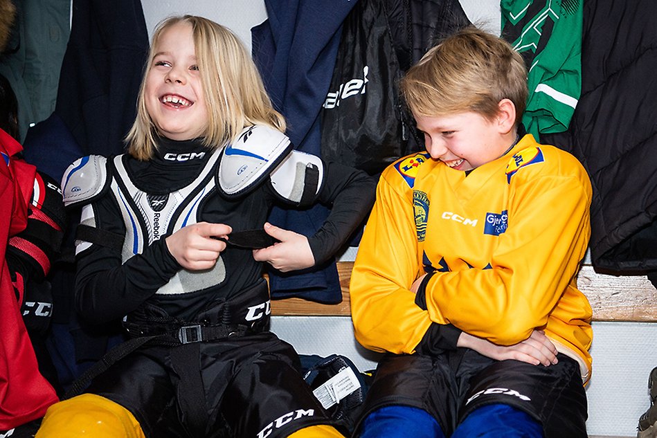 Två barn i hockeyutrustning sitter i omklädningsrummet och skrattar. Foto.