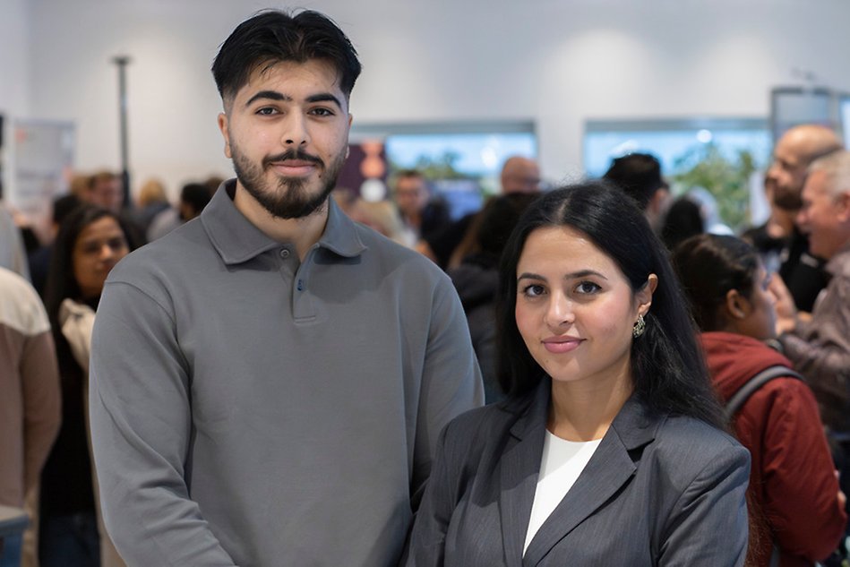 Two students in black hair and grey clothes are standing next to each other and are smiling to the camera. It's crowded in the background. Photo.