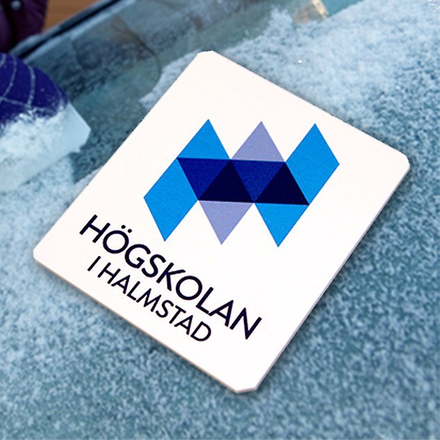 A white ice scraper with the Halmstad University logo in Swedish lies on a background of ice. Photo.