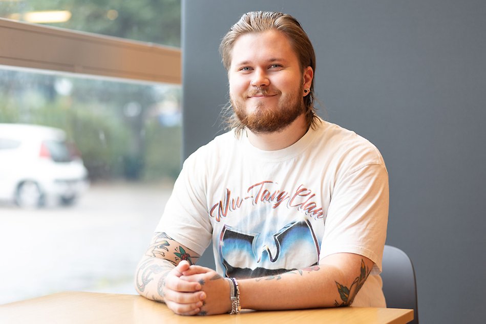 A young man with a beard is sitting at a table by a window. He looks at the camera and smiles. Photo.