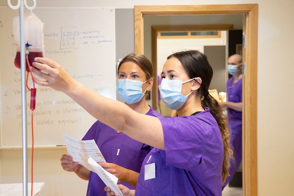 Två studenter i lila sjuksköterskekläder tittar på en blodpåse. Foto.