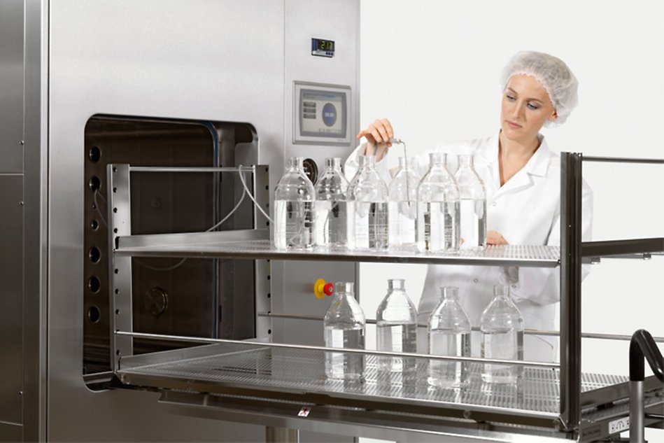 A woman in a white lab coat and hair cap is standing behind a steriliser with glass bottles. Photo.