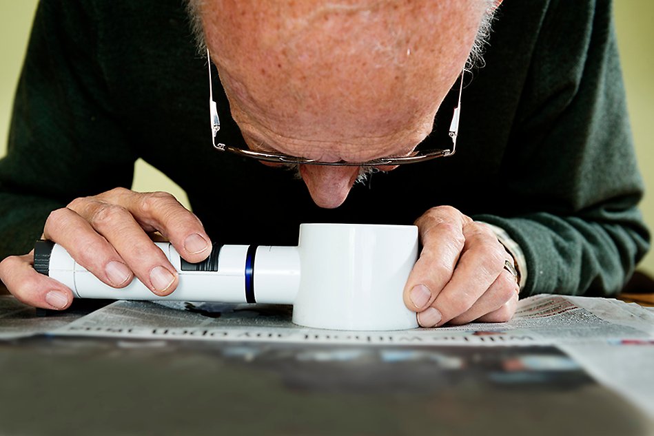 En äldre man sitter djupt böjd över ett förstoringsglas som han håll i med båda händerna, och läser en dagstidning som ligger på ett bord.