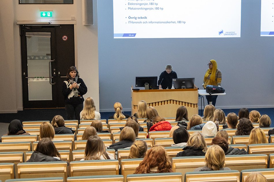 Tre kvinnliga forskare står på scen och presenterar i en sal. I publiken sitter unga kvinnor och lyssnar. Foto.
