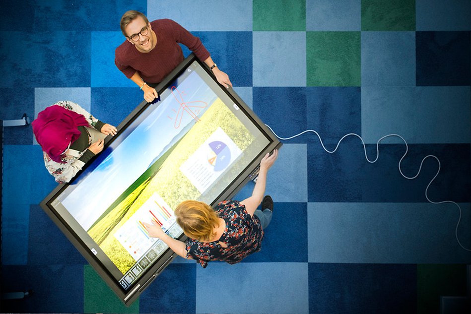 From above view of two women and one man are standing around a large, horizontal digital screen, showing charts and statistics. Photo.