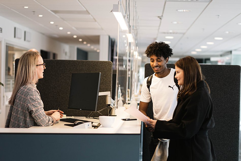 A person standing behind an information desk interacting with two people on the other side of the desk. Photo.