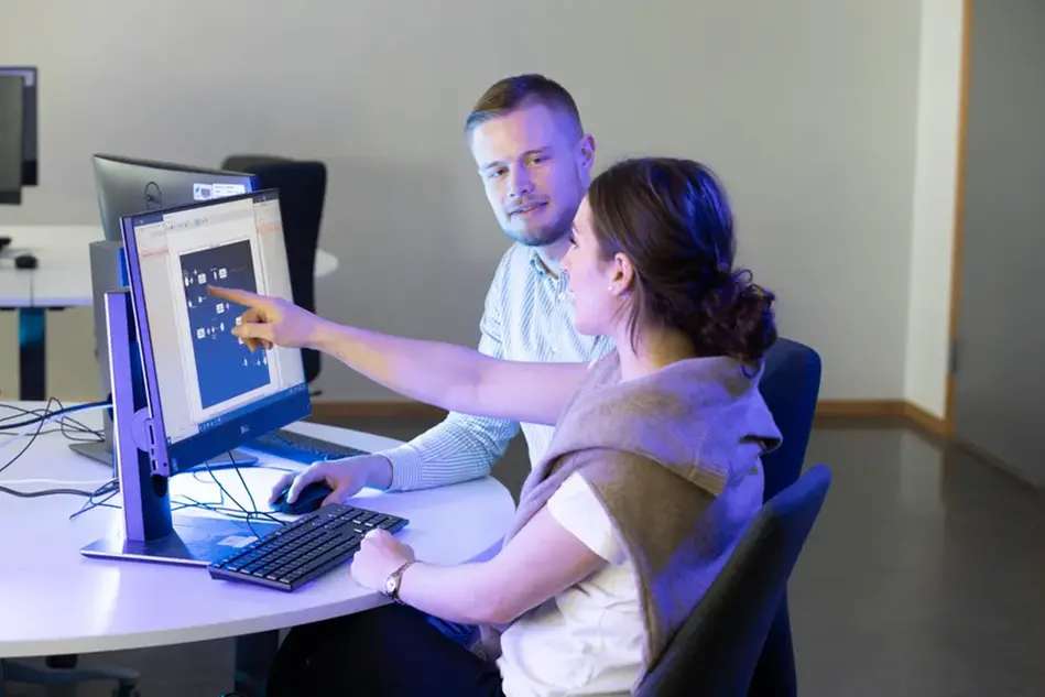 A woman and a man sit at a computer. The woman is pointing at the screen. Photo.