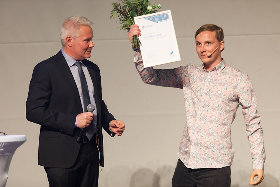 Two men standing in the spotlight, one of them holding up flowers and a diploma. Photo.