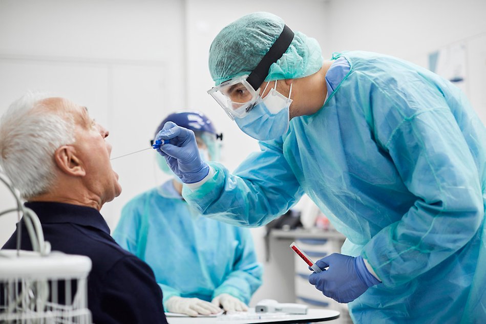 A person wearing protective medical clothing is swabbing the inside of an older man’s mouth. Photo.
