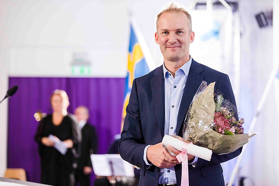Man iklädd kostym håller blombukett, diplom och ett äpple i glas, tittar in i kameran och ler. Foto.