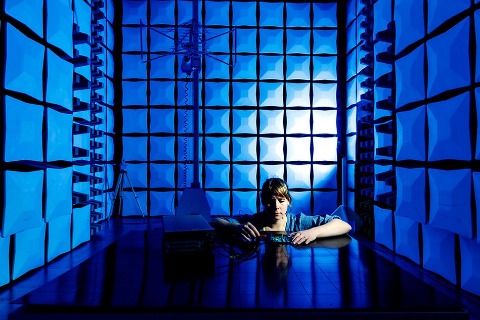 Kvinna sitter vid bord i testkammare för elektronik och håller i ett föremål, blått ljus i rummet. Foto.