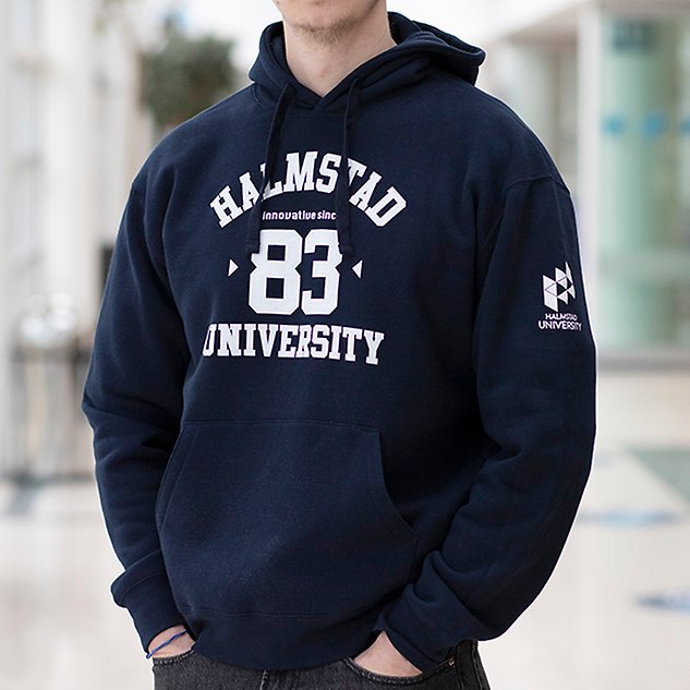 En mörkblå långärmad tröja med texten ”Högskolan 83 Halmstad” i vit text sitter på en person vars överkropp syns i bild. Foto.