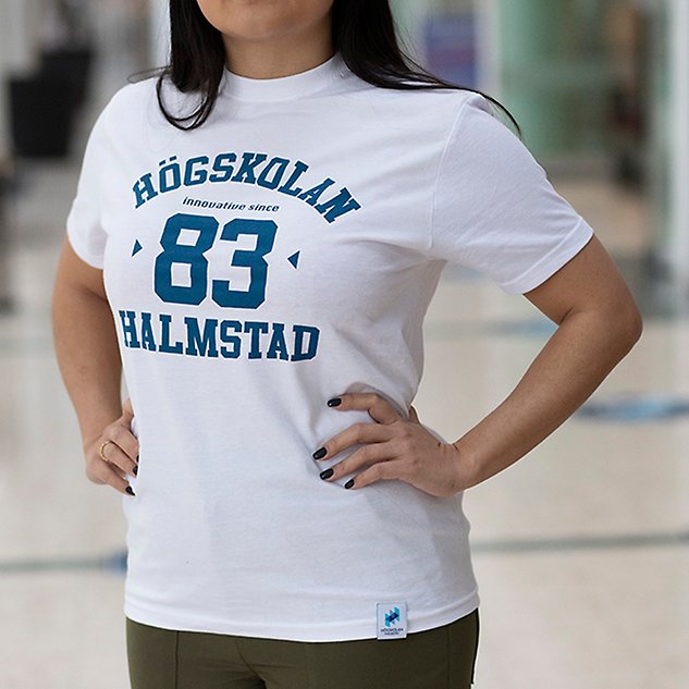 A white t-shirt with the text "Högskolan 83 Halmstad" in blue text is shown on a person whose upper body is visible in the picture. Photo.