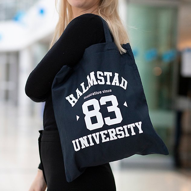 En mörkblå tygkasse med texten ”Halmstad 83 University” hänger på axeln på en person sedd från sidan. Foto.