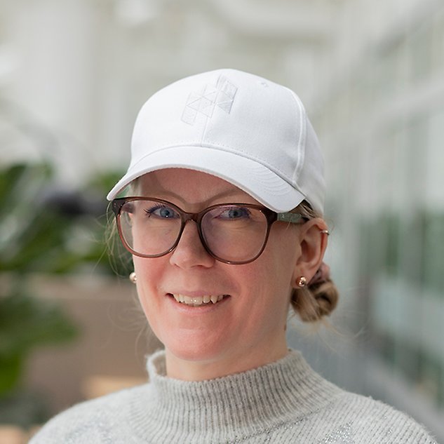 A smiling person with glasses is looking into the camera and has a white cap on her head. Photo.