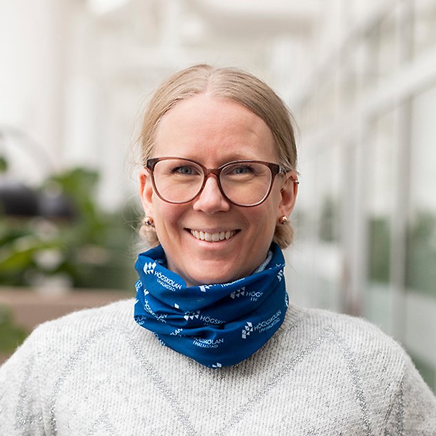 A smiling person with glasses is looking into the camera and has a blue scarf around her neck. Photo.