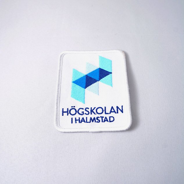 A white embroidered badge with the Halmstad University's Swedish logo on a white background. Photo.