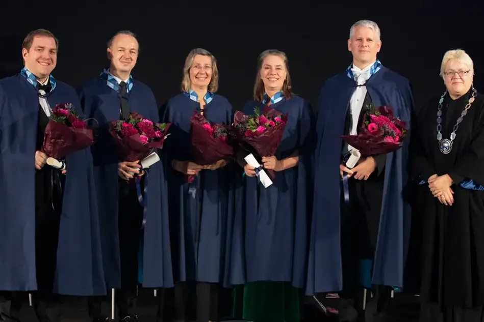 Fem personer i blå caper står uppradade på en scen. I famnen har de blommor och diplom. Lägst till höger står en person i svart rektorskappa och med en stor silverkedja runt halsen Foto.