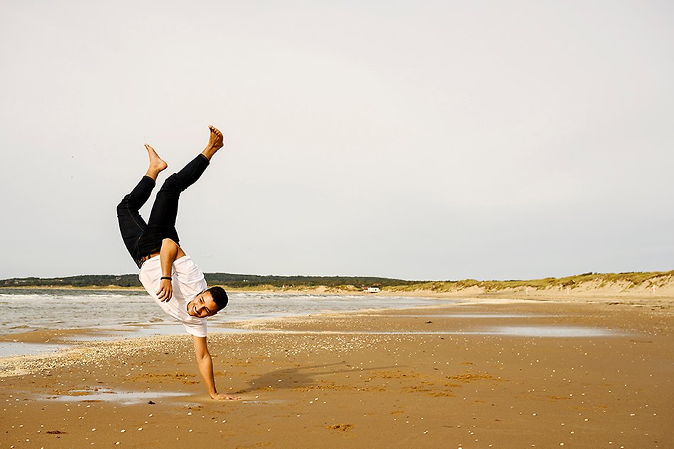 En glad, ung man i svarta byxor och vit t-shirt står på en hand i vattenbrynet på en strand. Foto.