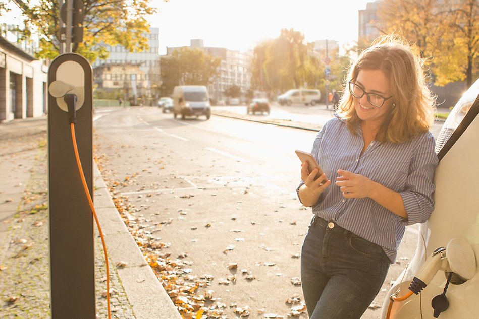 Photo of a woman charging her electric car while looking at her phone.