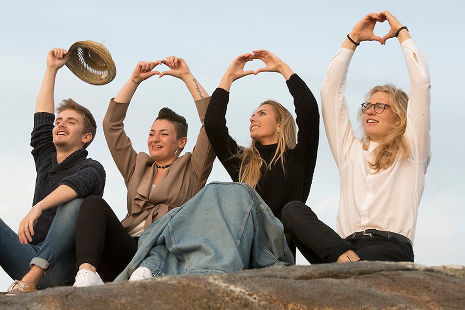 Fyra glada personer sitter på en klippa med solnedgångsljus i ansiktet och formar sina händer till var sitt hjärta. Foto.