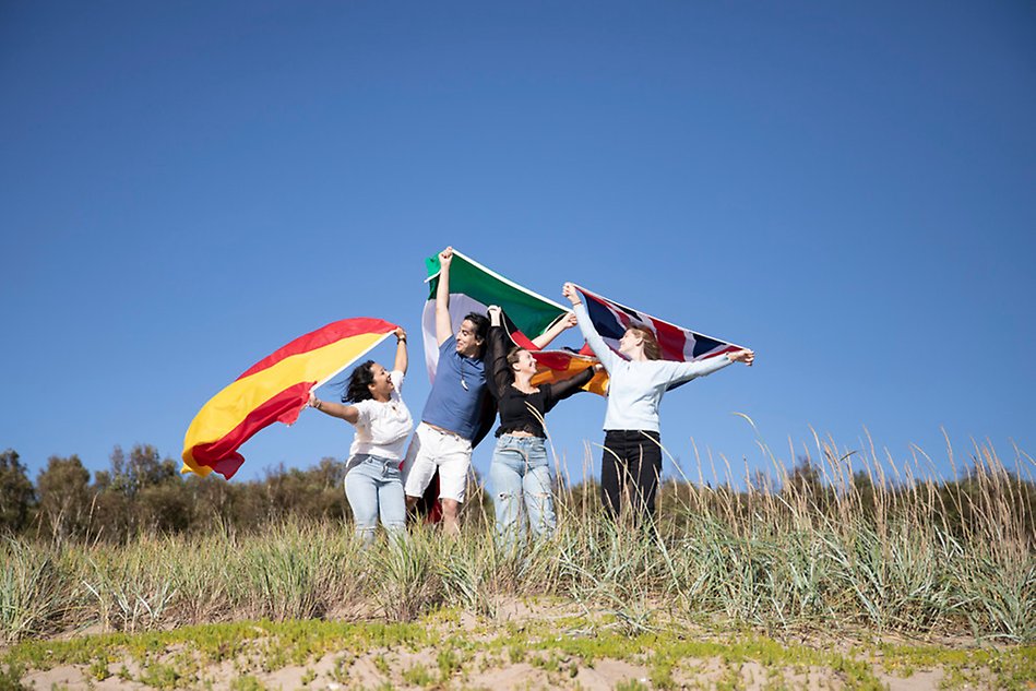 Fyra studenter står på stranden och håller upp olika landsflaggor ovanför huvudet. Foto.
