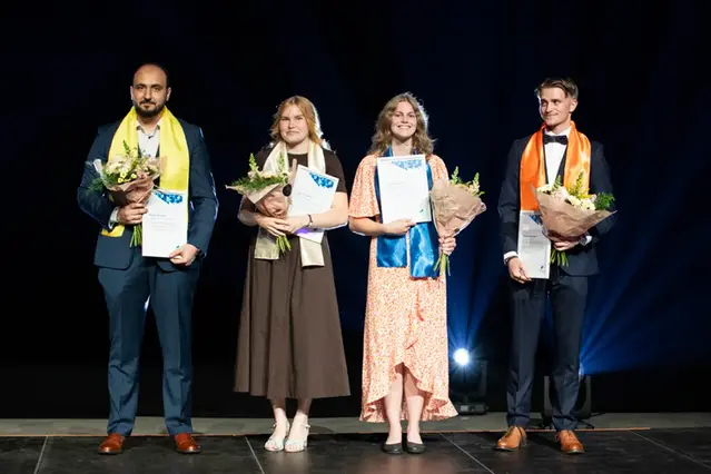 Four young people are standing on a stage in a row with flowers and diplomas in their hands.