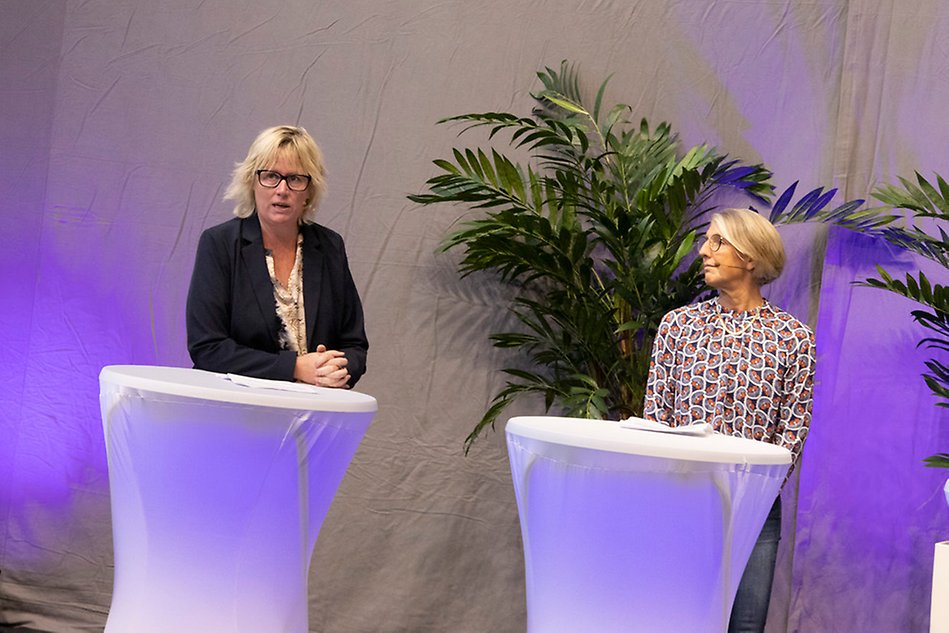 Two women wearing microphones standing by tables talking. Photo