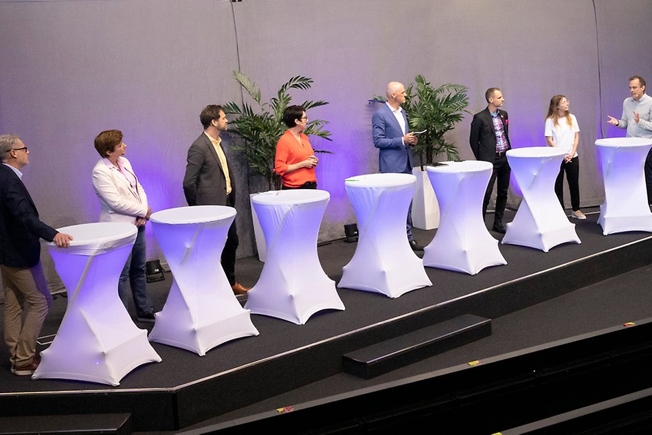 Seven people standing by tables looking at an eighth person talking. Photo.