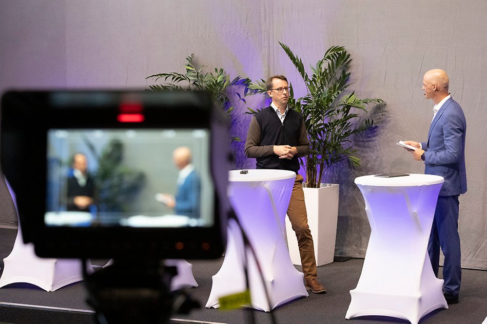 Two men standing by tables talking in focus and a camera showing the same thing out of focus. Photo.