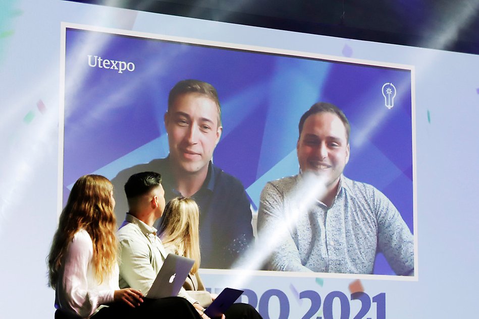 Bild på två personer på en stor skärm i bakgrunden. Till vänster sitter tre personer bortvända från kameran och tittar på personerna på skärmen.