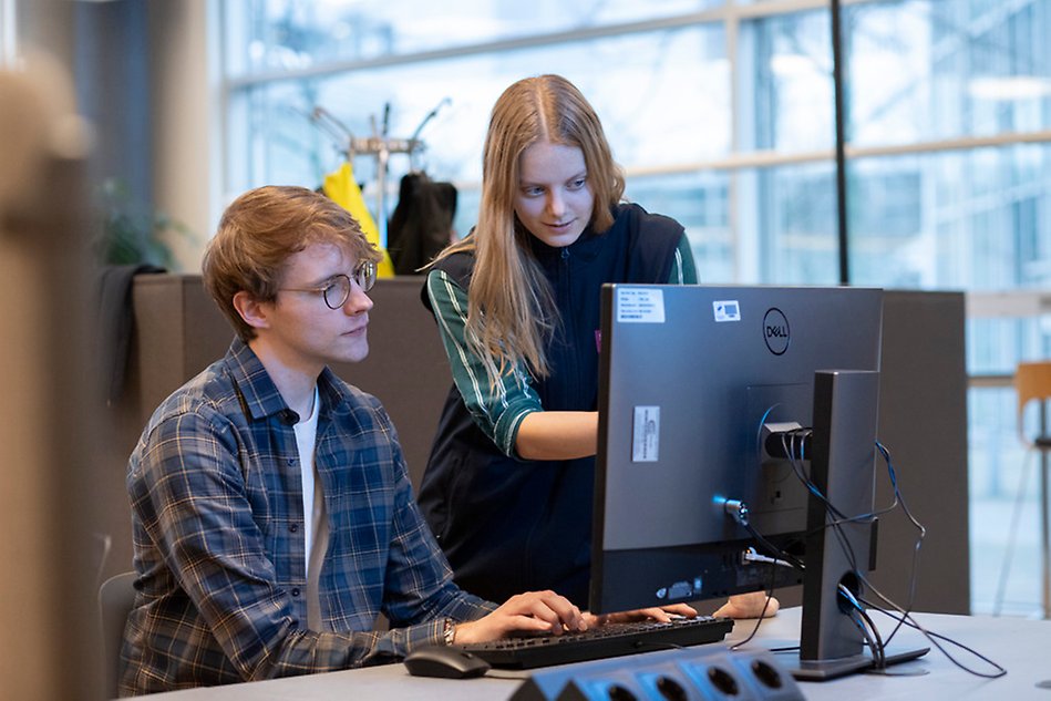 Two young people at a computer screen. One stands up and helps the person sitting down with something on the screen. Photo.