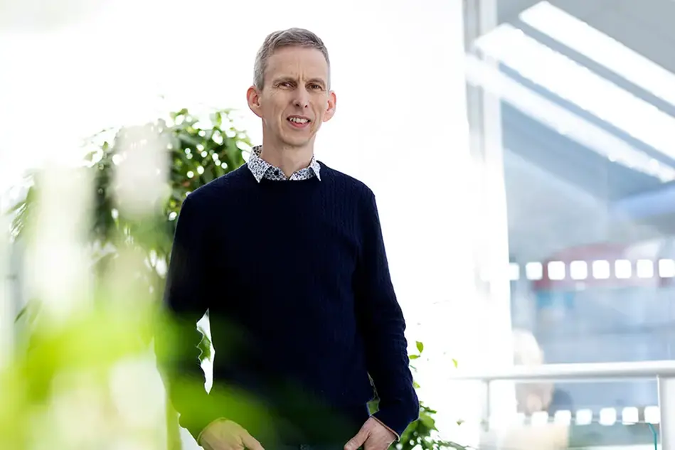 A man stands infront of a light background and looks into the camera. A plant is seen in the foreground.
