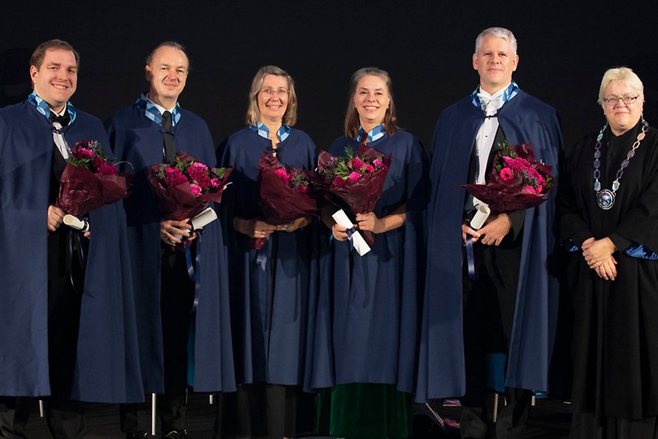 Fem personer i blå caper står uppradade på en scen. I famnen har de blommor och diplom. Lägst till höger står en  person i svart rektorskappa och med en stor silverkedja runt halsen Foto.