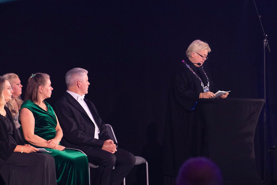 En korthårig kvinna i en svart käp står vid ett podium på en scen och håller ett tal. På scenen sitter en rad med uppklädda människor.