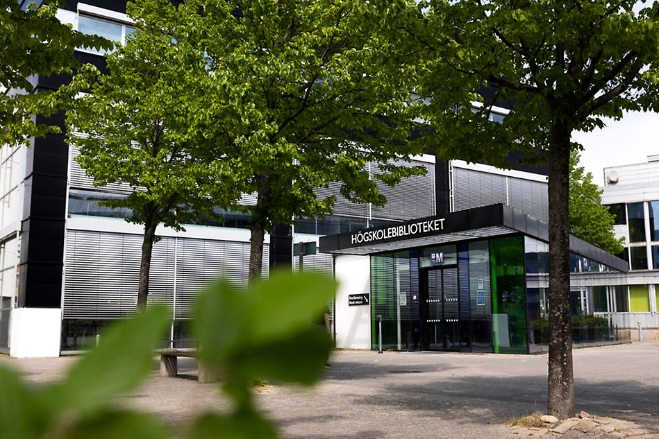 En byggnad med texten Högskolebiblioteket. Grön växtligthet i förgrunden.