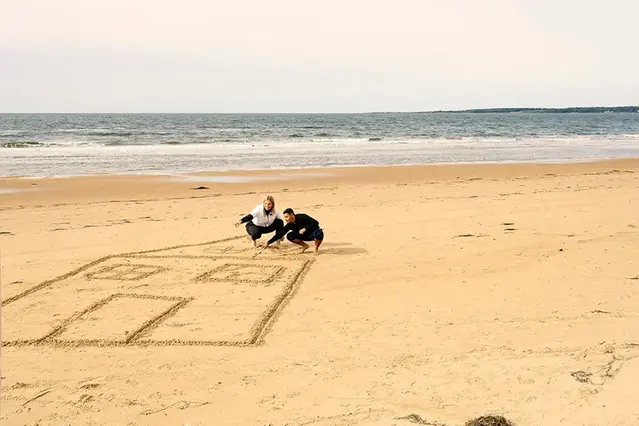 Två personer ritar ett stort hus i sanden på en strand. I bakgrunden syns havet. Foto.
