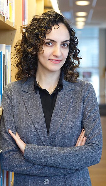  A smiling woman with curly dark hair and folded arms leaning against a bookcase. Photo.